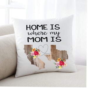 Könnte beinhalten: Ein weißes Kissen mit dem Text "HOME IS where my MOM IS" in schwarzen Buchstaben. Das Kissen zeigt ein Herz mit einer Linie, die zwei Holz-Staatsumrisse mit floralen Akzenten verbindet.