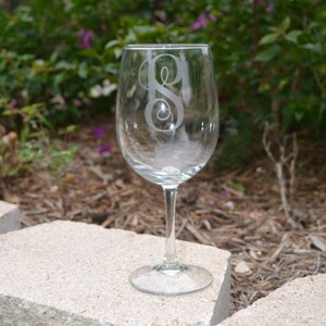May include: A clear glass wine glass with a single initial etched on the front. The glass is sitting on a light gray stone surface.