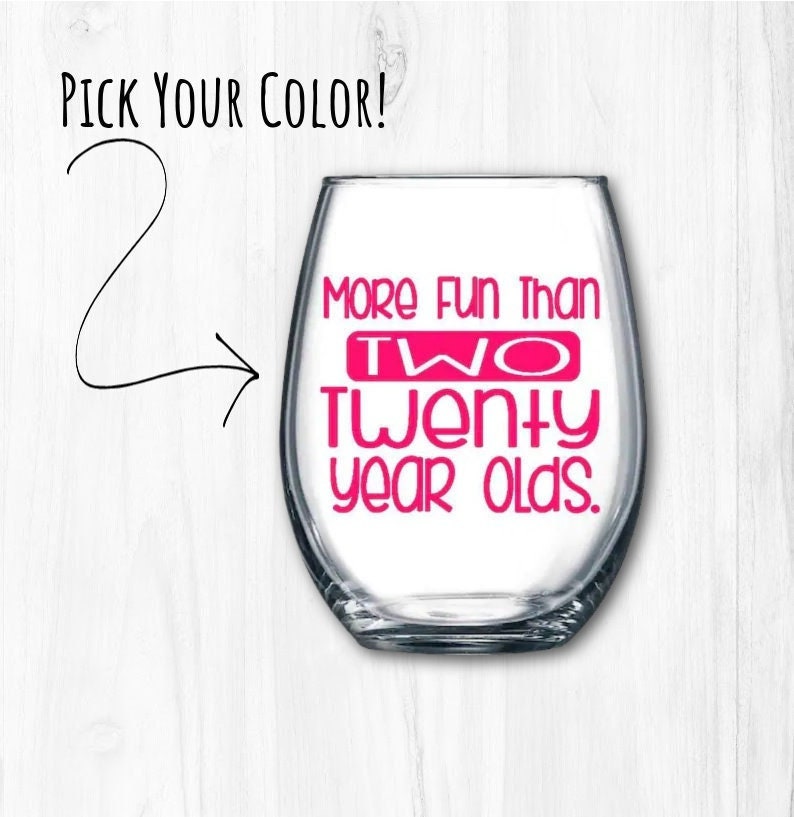 May include: A clear glass wine glass with the text "More Fun Than Two Twenty Year Olds." printed in pink.