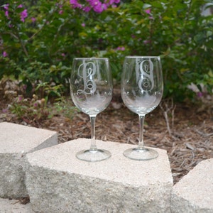 May include: Two clear glass wine glasses with etched initials on the front. The glasses are sitting on a gray stone surface.