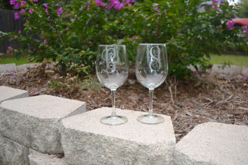 May include: Two clear glass wine glasses with etched initials on the front. The glasses are sitting on a gray stone surface.