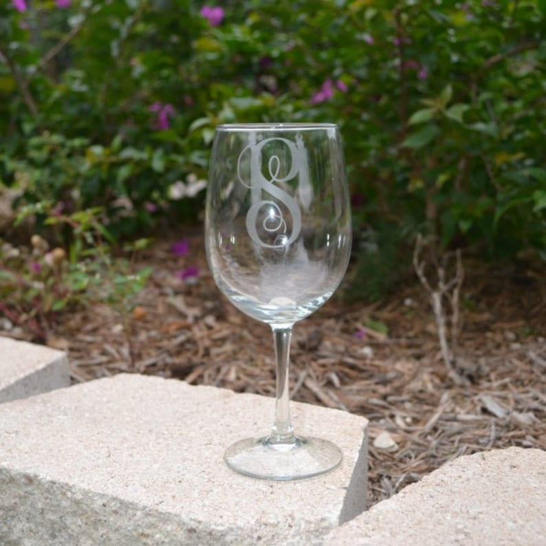 May include: A clear glass wine glass with a single initial etched on the front. The glass is sitting on a light gray stone surface.