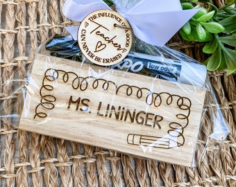 May include: A wooden gift tag with the text "The influence of a good teacher can never be erased" and a heart. The tag is tied with a white ribbon to a small wooden block with the text "Ms. Lininger" and a pencil illustration.