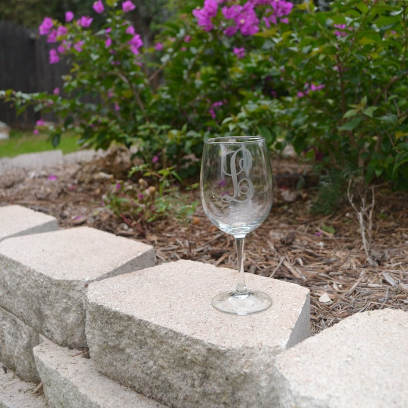 May include: A clear glass wine glass with a single initial etched into the side. The glass is sitting on a gray stone surface.
