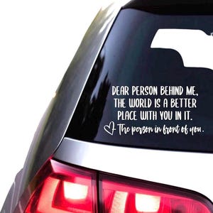 Op de afbeelding: Een witte vinyl sticker aangebracht op de achterruit van een voertuig. De tekst luidt: DEAR PERSON BEHIND ME, THE WORLD IS A BETTER PLACE WITH YOU IN IT. Het bevat een klein hartje en de handgeschreven tekst: The person in front of you.