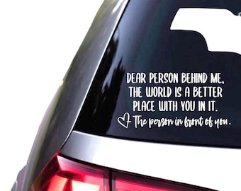 May include: A white vinyl decal applied to the rear window of a vehicle. The text reads: DEAR PERSON BEHIND ME, THE WORLD IS A BETTER PLACE WITH YOU IN IT. It includes a small heart icon and the script text: The person in front of you.