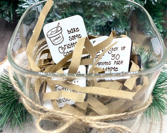Puede incluir: Un tarro de vidrio lleno de pequeños cuadrados de madera, cada uno con una actividad diferente con temática navideña escrita en él. Las actividades incluyen "hornear algunas galletas de Navidad", "quedarse despierto 30 minutos después de la hora de acostarse" y "escuchar música navideña".