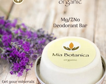 May include: A round white Mía Botanica organic Mg/ZNo deodorant bar in a metal tin, featuring a label with a logo of hands holding a flame. Beside the tin is a sprig of purple lavender. Text reads: Mía Botanica organic, Mg/ZNo Deodorant Bar, Get your minerals & smell great!
