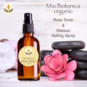 May include: A brown glass bottle with a black spray nozzle and a label that reads "Mia Botanica Rose Toner organic". The bottle is sitting on a white wooden surface with black stones and a pink flower.