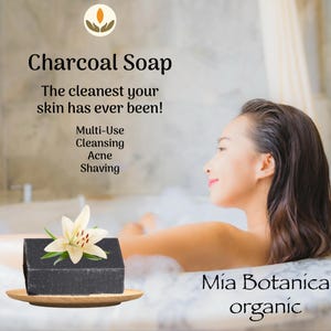 May include: A black charcoal soap bar with a white flower and green leaves on a wooden plate. The text on the image says "Charcoal Soap" and "The cleanest your skin has ever been!" with the words "Multi-Use, Cleansing, Acne, Shaving". The brand name "Mía Botanica organic" is also visible.