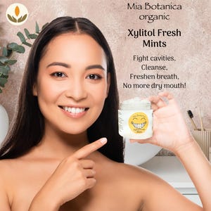 May include: A woman smiles, pointing to a small jar of Xylitol Fresh Mints. The jar has a yellow label with a smiling face and the text "Mia Botanica". The text on the image promotes the mints for fighting cavities, cleansing, freshening breath, and eliminating dry mouth.