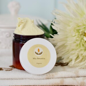 Op de afbeelding: Een pot gele cr&egrave;me met het deksel eraf, waardoor het product zichtbaar is. Het deksel is wit met een geel etiket met de tekst "Mia Botanica Organic". Een witte bloem en groene bladeren zijn op de achtergrond.