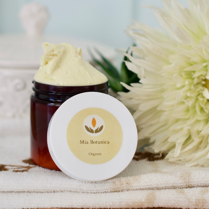 Op de afbeelding: Een pot gele cr&egrave;me met het deksel eraf, waardoor het product zichtbaar is. Het deksel is wit met een geel etiket met de tekst "Mia Botanica Organic". Een witte bloem en groene bladeren zijn op de achtergrond.