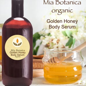 May include: A dark brown bottle of Mia Botanica Golden Honey Body Serum with a black cap. A small glass jar with honey and a wooden honey dipper is next to the bottle. The words "Mia Botanica organic Golden Honey Body Serum" are printed on the bottle.