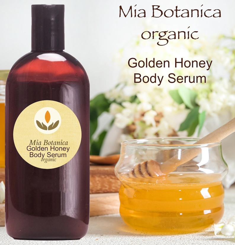 May include: A dark brown bottle of Mia Botanica Golden Honey Body Serum with a black cap. A small glass jar with honey and a wooden honey dipper is next to the bottle. The words "Mia Botanica organic Golden Honey Body Serum" are printed on the bottle.