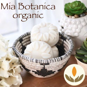 May include: Three white organic soap bars in a black and white woven basket. The soap bars are shaped like flowers with a raised design. The text "Mia Botanica organic" is visible in the background.