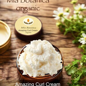 Op de afbeelding: Een pot witte Amazing Curl Cream met de tekst "Amazing Curl Cream" onderaan. Het product is van Mia Botanica, zoals aangegeven op het etiket. Op het etiket staat ook "organic". De achtergrond is een houten oppervlak met bloemen.