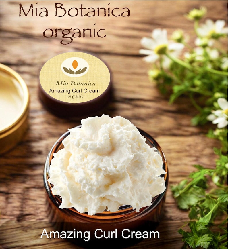Op de afbeelding: Een pot witte Amazing Curl Cream met de tekst "Amazing Curl Cream" onderaan. Het product is van Mia Botanica, zoals aangegeven op het etiket. Op het etiket staat ook "organic". De achtergrond is een houten oppervlak met bloemen.