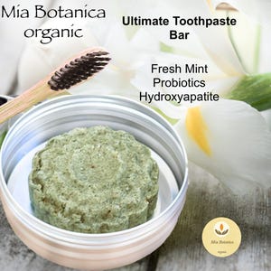 May include: A green toothpaste bar in a silver tin with a wooden toothbrush and a white flower in the background. The text on the label reads "Mia Botanica organic Ultimate Toothpaste Bar Fresh Mint Probiotics Hydroxyapatite".
