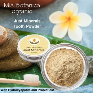 May include: A small, white jar of Mia Botanica Just Minerals organic tooth powder. The jar is open and the powder is visible. The powder is a light brown color. The jar is sitting on a brown rock. The background is a green leaf and a white flower.
