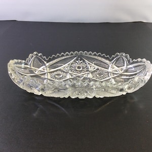 May include: A clear glass serving dish with a scalloped edge and intricate cut-glass design. The dish is oval shaped and has a raised center.