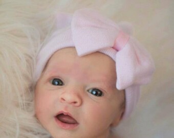 May include: A newborn baby wearing a pink bow headband with a white fuzzy background.
