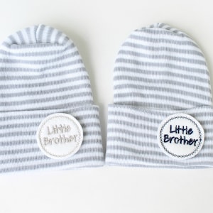 May include: Two gray and white striped baby hats with white circles that say "Little Brother" in different colored embroidery.