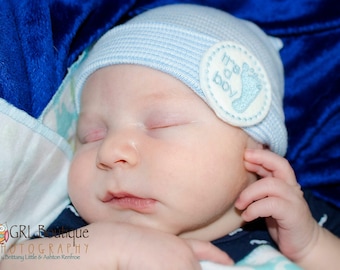 May include: A sleeping baby wearing a light blue knitted hat with a white embroidered foot print and the text "It's a boy".