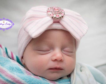 May include: A newborn baby wearing a pink and white striped knit hat with a large, round, pink jeweled embellishment. The baby is wrapped in a blue, pink, and white striped blanket.