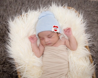 May include: A newborn baby wearing a blue and white striped hat with a red heart patch that says "You" is sleeping on a white fluffy blanket.