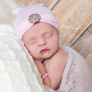 May include: A sleeping newborn baby wearing a pink knit hat with a jeweled embellishment. The baby is wrapped in a white crocheted blanket.