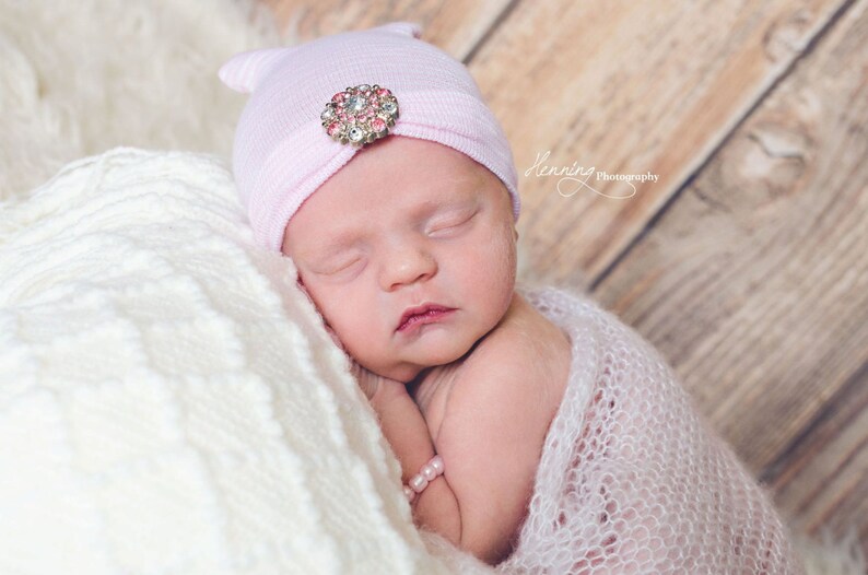 May include: A sleeping newborn baby wearing a pink knit hat with a jeweled embellishment. The baby is wrapped in a white crocheted blanket.