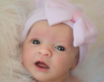 May include: A baby wearing a pink knit hat with a bow on top. The hat is made of soft, stretchy fabric and has a comfortable fit.
