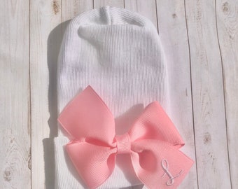 May include: White knitted baby hat with a pink bow and the letter "L" embroidered on the bow.