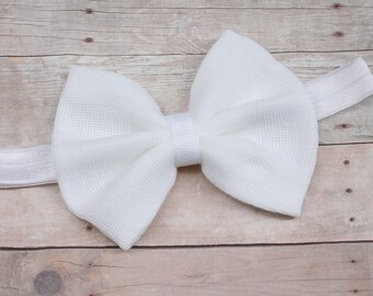 May include: A white baby headband featuring a large bow made of textured fabric. The bow is centered on a matching white elastic band. The headband is designed for infants and toddlers.