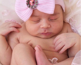 May include: A newborn baby wearing a light purple knit hat with a pink striped bow and a sparkling pink jewel. The baby is nestled in soft, light-colored fabric. The text "Quick Ship" is visible.