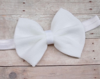 May include: A white fabric bow headband. The bow is tied in a large, fluffy knot and is attached to a white elastic headband.