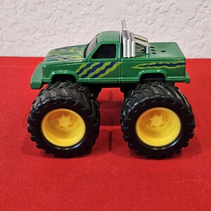 May include: A green monster truck toy with large black tires and yellow rims. The truck has a green body with blue and yellow patterned accents. It features a roll bar and exhaust pipes.