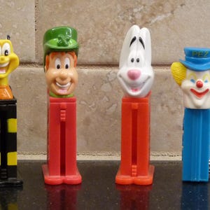 May include: Four colorful PEZ dispensers. A yellow bee, a leprechaun with a green hat, a white rabbit, and a clown with a blue hat. The dispensers have red, black, and blue bases. The clown has a silver ring attached.