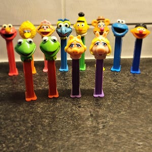 May include: A collection of colorful PEZ dispensers featuring various characters. The dispensers include characters from Sesame Street and the Muppets, with vibrant colors and detailed character heads. The dispensers are arranged on a dark surface.
