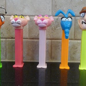 May include: A collection of five colorful PEZ dispensers featuring cartoon characters. The dispensers include an orange ant, two pink panthers, a blue aardvark, and a detective. Each dispenser has a rectangular base and a character head.