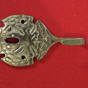 May include: A decorative metal wall hook with a unique design. The hook features a stylized, openwork pattern with a central oval and a small hook at the end of a handle. The metal has a silver finish.