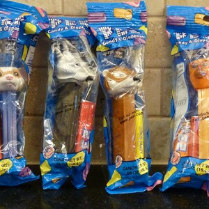 May include: Four PEZ candy dispensers in their original packaging. The dispensers feature animal heads: a dog, a wolf, a fox, and a lion. Each package is blue with the words "Candy & Dispenser" printed on it.