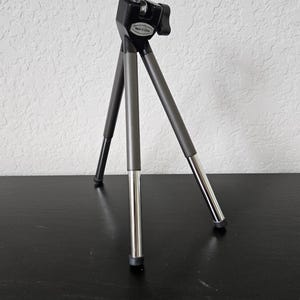 May include: A small, black and gray tripod with three legs. The legs are a combination of gray and silver metal. The top has a black mounting plate and a screw for attaching a camera or phone. The tripod is designed to be compact and portable.