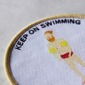 May include: A white embroidered patch with a tan border. The patch features a cartoon illustration of a person wearing swimming trunks and arm floaties. The text "KEEP ON SWIMMING" is written in black around the edge of the patch.