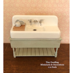 May include: White porcelain farmhouse sink with two chrome faucets and a wooden dish rack with white dishes. The sink is mounted on a light blue cabinet with a white shelf.