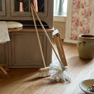 Może przedstawiać: Miniaturowy zestaw drewnianego miotły i mopa z białymi szczecinami i białą bawełnianą głowicą mopa. Zestaw znajduje się na drewnianej podłodze w otoczeniu domku dla lalek. Tiny Dwelling Miniatures & Decorations 1:12 Scale.