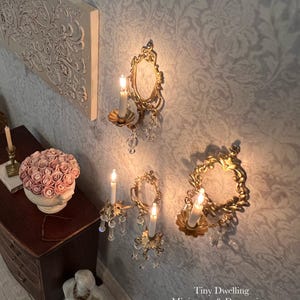 May include: Miniature gold-colored wall sconces with lit candles and crystal accents. The sconces are mounted on a patterned wall. A small wooden dresser and a decorative floral arrangement are also visible.