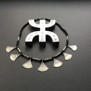 May include: A silver necklace with a black beaded chain and fan-shaped pendants. A matching silver pendant with etched designs is positioned behind the necklace. The jewellery is displayed on a dark grey surface.