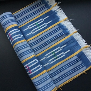 May include: Rolled blue and white striped fabric with yellow and green accents. The fabric has a fringed edge and is likely used for clothing or home decor. The design includes white and yellow horizontal stripes.